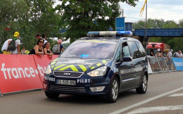 Polizeiauto fährt an einer Menge vorbei, die sich an einem Geländer mit Bannern befindet, mit Bäumen, einer Brücke und einer Flagge im Hintergrund.