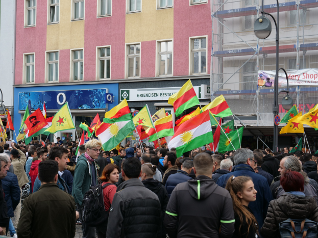 Eine große Gruppe von Menschen geht eine Straße entlang und hält kurdische Flaggen in den Händen, während Laternenmasten und Gebäude mit Fenstern die Straße säumen und Texttafeln im Hintergrund zu sehen sind.