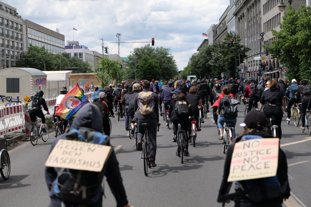 Eine große Gruppe von Menschen, die auf Fahrrädern eine Straße entlangfahren, einige halten Schilder, mit einem Zaun auf der linken Seite, Bäumen, Gebäuden, Laternen und Verkehrszeichen im Hintergrund.