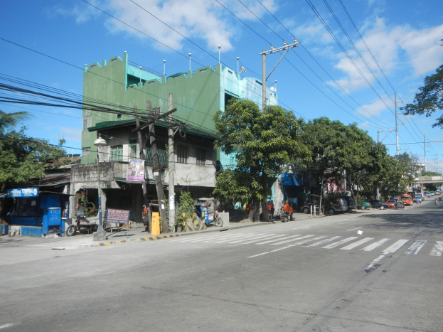 Eine Stadtstraße mit einem grünen Gebäude, fahrenden Fahrzeugen, Fußgängern auf dem Gehweg, Stromleitungen und einem bewölkten Himmel.