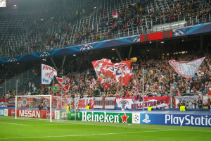 Fußballspiel in einem hell erleuchteten Stadion mit einer großen Zuschauermenge, grünem Rasen, Tor, Bannern und einem Bildschirm in der linken oberen Ecke.