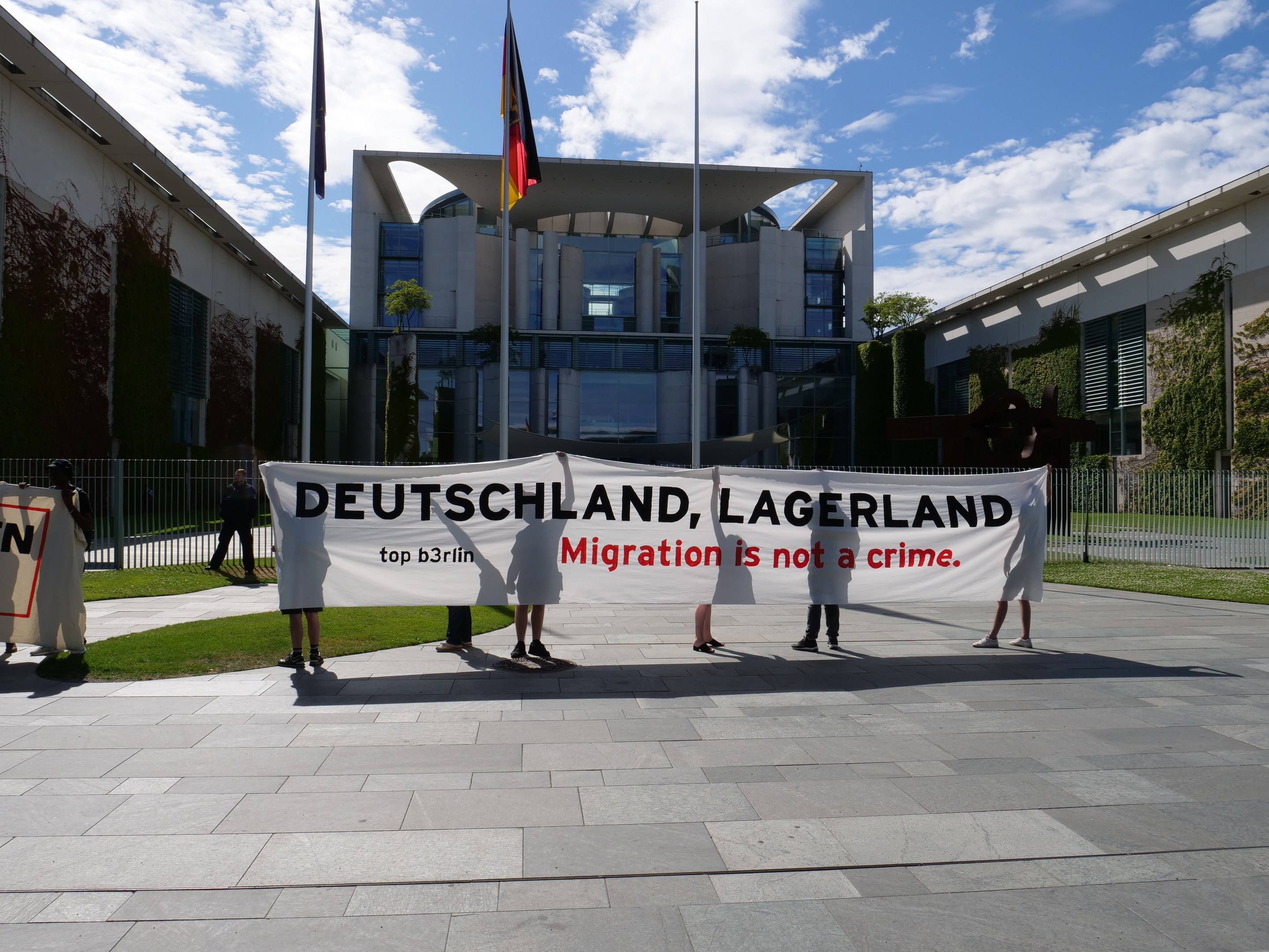 Gruppe von Menschen, die eine Tafel mit der Aufschrift "Deutschland, Lagerland Migration ist kein Verbrechen" vor einem Fenster mit Fenstern, umgeben von Gras, einem Metallzaun, Pflanzen, Bäumen und Fahnen mit Stäben unter einem bewölkten Himmel halten.