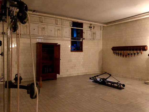 Ein unordentlicher Raum im ehemaligen Gefängnis St. Louis mit Seilen, Fitnessgeräten und anderem Equipment, einem Schrank, einem Fenster und einer Deckenlampe im Hintergrund.