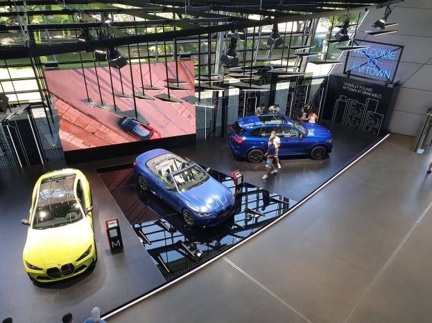 Eine Gruppe von Menschen im BMW Museum in München, Deutschland, mit Autos zur Linken und einem Schild mit der Aufschrift "BMW Museum München" zur Rechten, mit Blick auf eine Außenszene mit Bäumen, Gebäuden und Himmel durch Glasfenster.