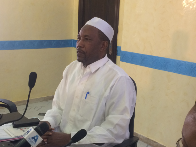 Ein Mann in weißem Anzug und Hut sitzt an einem Tisch mit Mikrofonen, spricht, während Papiere und Gegenstände auf dem Tisch zu sehen sind, mit einem anderen Mann mit Brille rechts von ihm, in einem Raum mit einer Wand und einer Tür im Hintergrund.