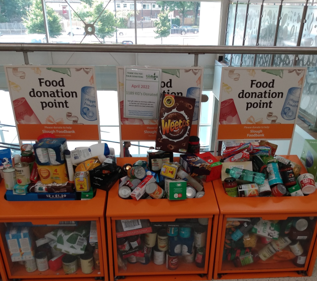 Orange Schübe mit Lebensmitteln wie Flaschen, Dosen und Kisten in einer Spendenstelle, mit Informationsschildern und einem Blick auf Fahrzeuge, Bäume, Gebäude und Himmel durch Glasfenster.