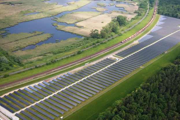 Eine Luftaufnahme eines Solarparks mit zahlreichen Solarmodulen auf einem Feld, umgeben von Bäumen, Gras und Wasser, mit einem Zug, der auf einem Bahngeleis in der Nähe fährt.