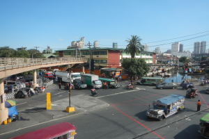 Eine belebte Stadtstraße mit Autos, Bussen und Motorrädern, eine Brücke mit Geländern und Säulen, Laternenmasten, Strommasten mit Drähten, Gebäude mit Fenstern, Bäume und ein bewölkter Himmel.
