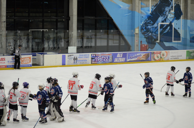 Junge Menschen in Helmen und Hockey-Ausrüstung spielen Eishockey auf einer Indoor-Eisfläche mit Bannern, Säulen und Glaswänden im Hintergrund.