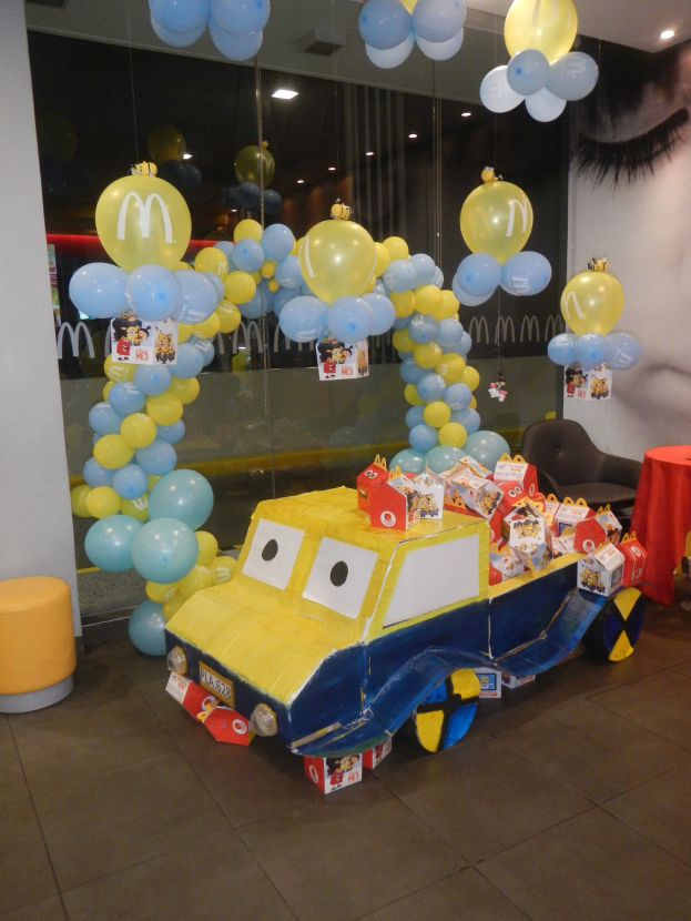 Eine McDonald's-Geburtstagsfeier mit einem gelben und blauen Lastwagen, der mit Ballons und anderen Dekorationen geschmückt ist, einem rot gedeckten Tisch mit Gegenständen, Hockern und einer Glaswand im Hintergrund und festlicher Deckenbeleuchtung.