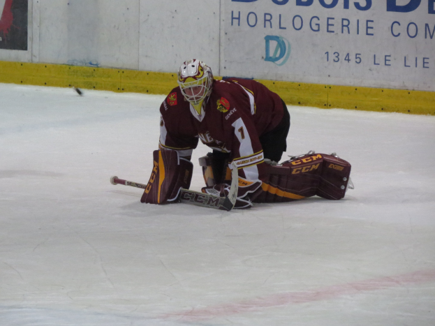 Eishockeyspieler in rot-gelber Uniform, der einen Schuss abwehrt, trägt Helm, Handschuhe und Knieprotektoren und hält einen Eishockeyschläger, mit einer Wand im Hintergrund, auf der etwas geschrieben steht.
