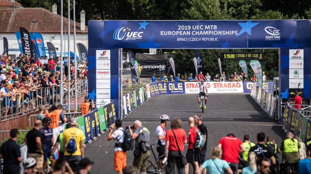Radfahrer, die unter einem Bogen mit der Aufschrift "2019 UEC MTB Europameisterschaften" eine Straße hinunterfahren, mit Zuschauern an den Seiten und Bäumen, Gebäuden und einem klaren blauen Himmel im Hintergrund.
