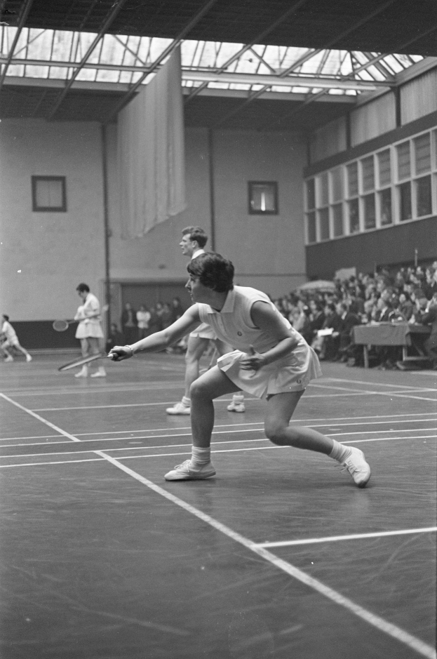 Schwarzes und weißes Foto von zwei Frauen, die Badminton auf einem Court spielen, jede hält einen Schläger, mit Zuschauern rechts und einer Wand mit Fenstern im Hintergrund.