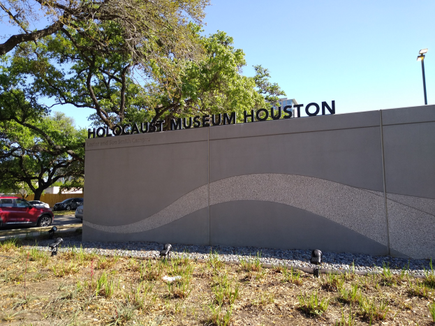 Außenansicht des Holocaust-Museums Houston mit eingravierter Textwand, Gedenksteine, Landschaft, Straßenverkehr, Laterne und bewölktem Himmel.