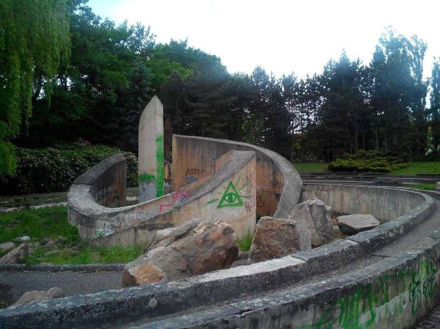 Verlassener Skatepark mit Graffiti an der Seite, umgeben von Felsen, Gras, Pflanzen und Bäumen unter einem klaren blauen Himmel.