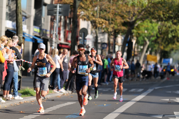 Eine Gruppe von Läufern bei einem Marathon auf einer Stadtstraße, mit Zuschauern auf der linken Seite, Bäumen und Gebäuden im Hintergrund unter einem klaren blauen Himmel, alle tragen Turnschuhe und Hüte.