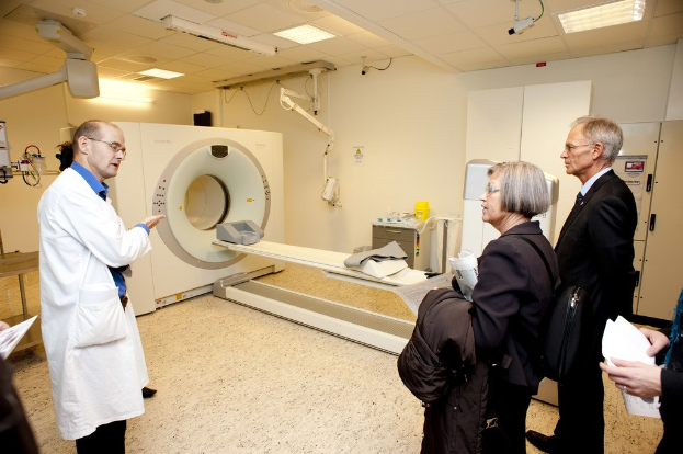 Arzt in weißem Kittel, der eine CT-Scan-Maschine zwei Personen in schwarzen Mänteln erklärt.