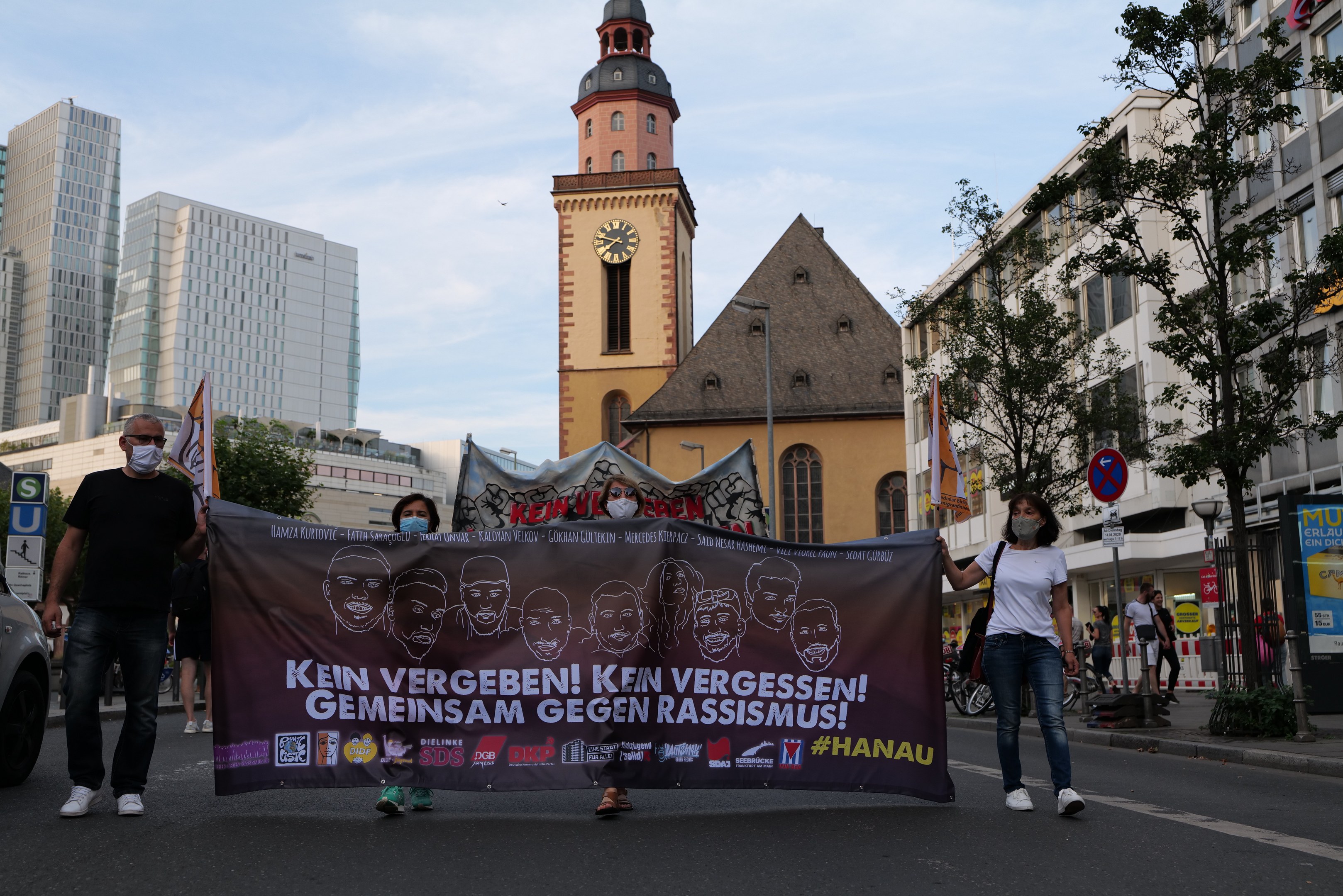 Eine Gruppe von maskierten Menschen marschiert mit einem Banner die Straße entlang, mit einem geparkten Auto auf der linken Seite und Gebäuden, Bäumen, Schildern, Pfählen und einem Kirchturm im Hintergrund unter einem klaren blauen Himmel.