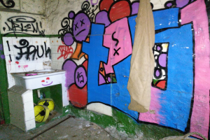 Badezimmer mit beschmierten Wänden, ein Waschbecken in der Ecke mit einem Tuch bedeckt und verstreute Gegenstände auf dem Boden.