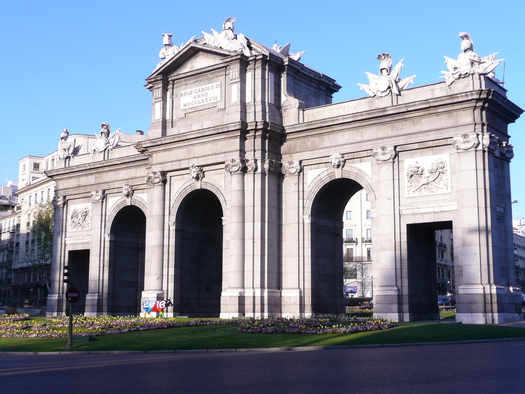 Gebäude und Skulpturen in der Mitte, mit einer Straße, Gras, Blumen, einem Pfahl und einer Tafel unten und Himmel oben.