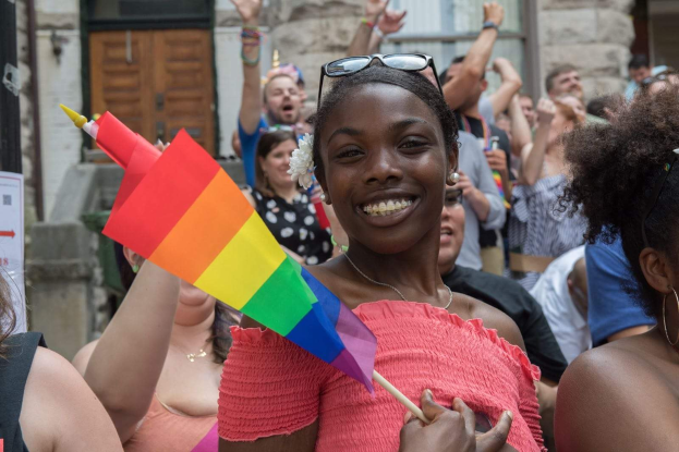 Eine Gruppe von Menschen vor einem Gebäude stehend, mit einer Frau in der Mitte, die eine Regenbogenfahne hält, wahrscheinlich an einem Christopher-Street-Day teilnehmend.