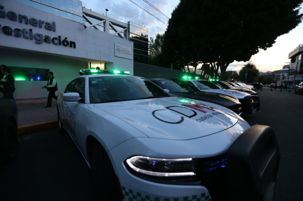 Eine Reihe von Polizeiwagen vor einem Gebäude mit Text geparkt, umgeben von Menschen, Bäumen, Laternenmästen, Drähten und einem bewölktem Himmel.