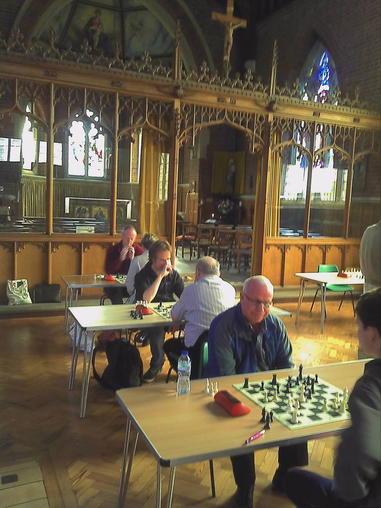 Eine Gruppe von Menschen, die an Tischen in einer Kirche Schach spielen, wobei ein Mann in einem blauen Hemd eine Flasche und einen Stift auf seinem Tisch hat, und ein Glasfenster mit einem Kreuzsymbol im Hintergrund.