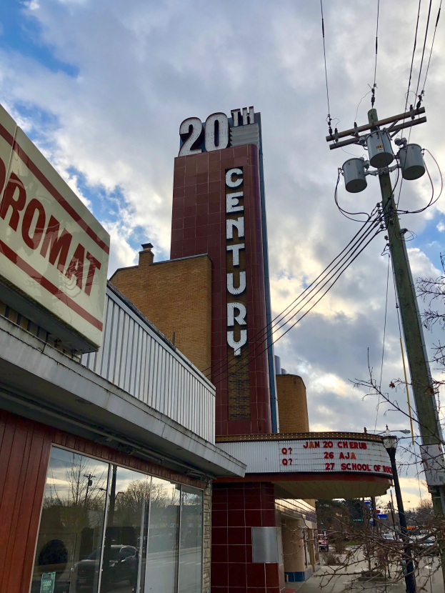 Eine Gruppe von Menschen steht in einem Cinemex-Kino in einem Einkaufszentrum, mit einer Person auf einem Stuhl, Tischen, Fotorahmen an der Wand, einem Schild, Deckenleuchten und Geräten auf den Tischen.