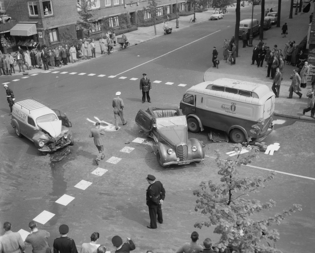 Schwarze und weiße Szene eines Autounfalls an einer Straßenkreuzung mit mehreren Fahrzeugen, darunter ein Van, und einer Gruppe von Zuschauern; Gebäude, Bäume, Pfosten und Texttafeln sind im Hintergrund sichtbar.