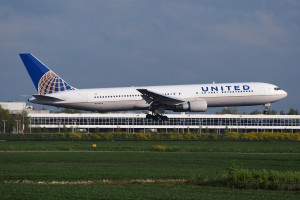 United Airlines Boeing 787-9 Dreamliner beim Abheben von einer Start- und Landebahn mit grünem Gras und Pflanzen, einem Gebäude und Bäumen im Hintergrund und einem klaren blauen Himmel.