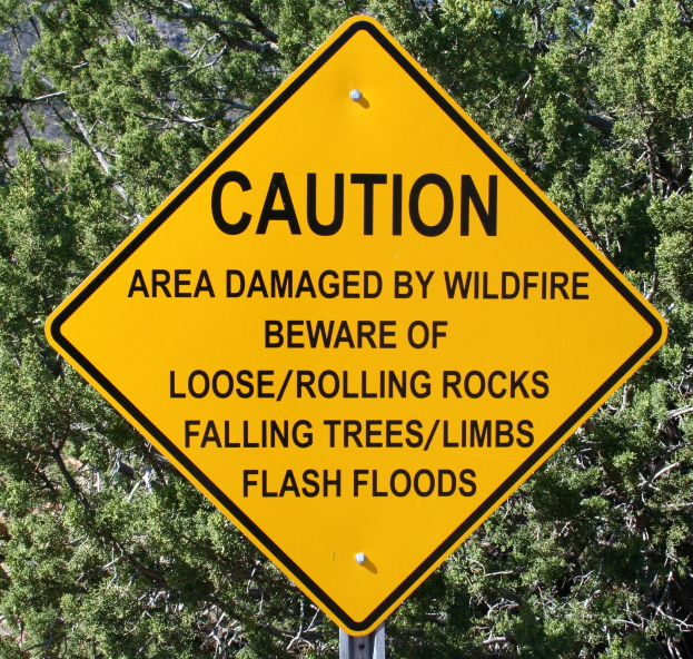 Gelbes Warnschild an einem Pfosten in der Nähe, das vor Wildfire-Schäden, lockeren Steinen, umstürzenden Bäumen und Überschwemmungen warnt, mit Bäumen im Hintergrund.