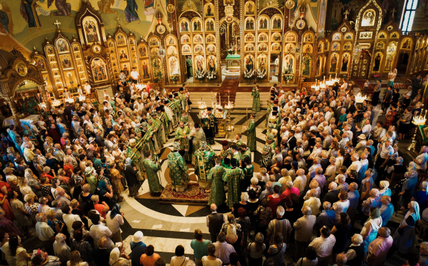 Eine große Gruppe von Menschen steht vor einer russisch-orthodoxen Kirche, einige halten Musikinstrumente, mit brennenden Kerzen, einem Teppich, Fotorahmen und einem Fenster.