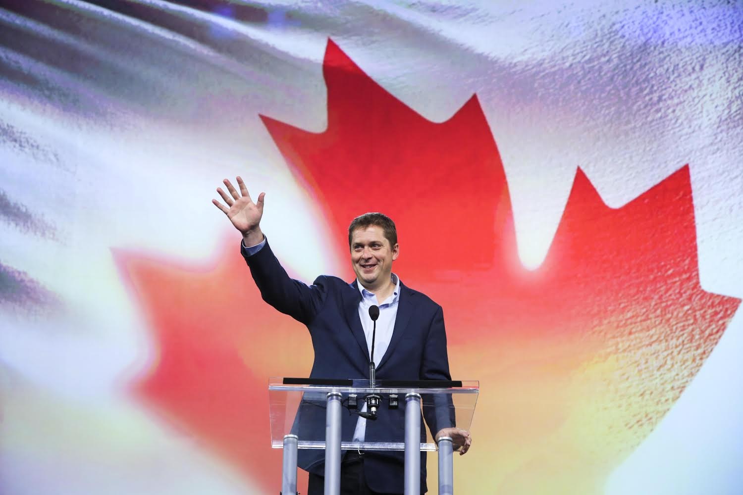 Der kanadische Premierminister Alex Salmond steht an einem Podium mit Mikrofonen, lächelt und schaut in die Kamera, vor einer kanadischen Flagge mit einem Bildschirm im Hintergrund.