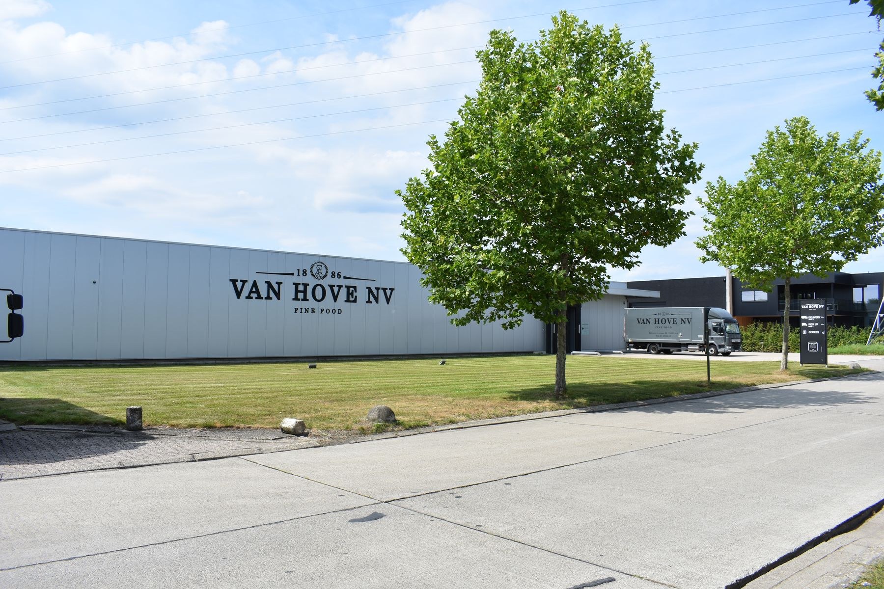 Eine Van Hove NV-Anlage in den Niederlanden mit Fahrzeugen, Gras, Bäumen, Gebäuden, Schildern, Pfählen und anderen Gegenständen im Vordergrund und einem bewölkten Himmel im Hintergrund.