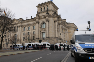 Eine Gruppe von Polizisten vor dem Reichstaggebäude in Berlin, Deutschland, mit Fahrzeugen, einem Zaun, Verkehrsampeln, Laternen, Bäumen und Flaggen im Hintergrund.