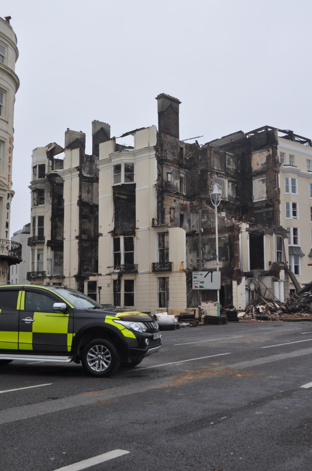 Ein Polizeiwagen vor einem stark beschädigten Gebäude mit zersplitterten Fenstern und verstreuter Trümmer, unter einem sichtbaren Himmel mit nahen Gebäuden.