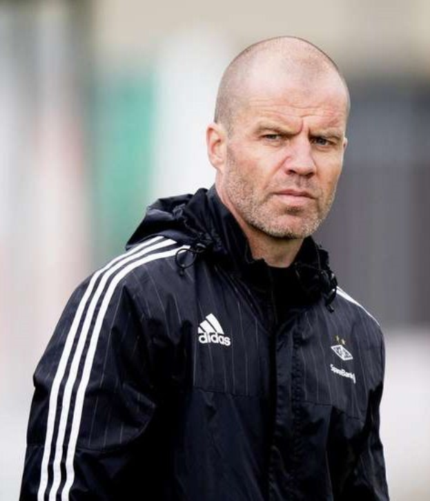 Baldiger Trainer in marineblauer Jacke mit weißen Streifen und weißem Kragen, der auf einem Fuballfeld steht und nachdenklich schaut.