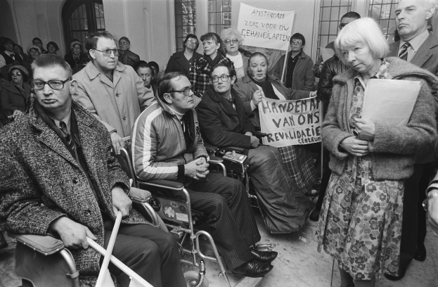 Gruppe von Menschen in Rollstühlen vor einer Menge, einige mit Schildern, mit Fenstern und einer Wand im Hintergrund.