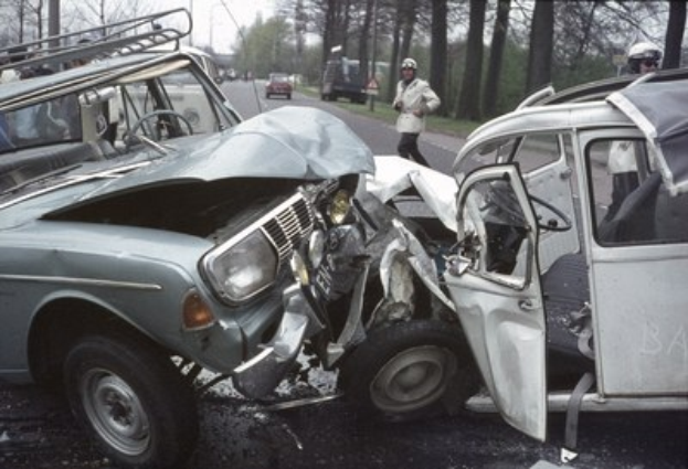 Ein beschädigt auf der Straße stehendes Auto mit Zuschauern und Einsatzkräften mit Helmen, vor Bäumen, Masten und einem klaren blauen Himmel.