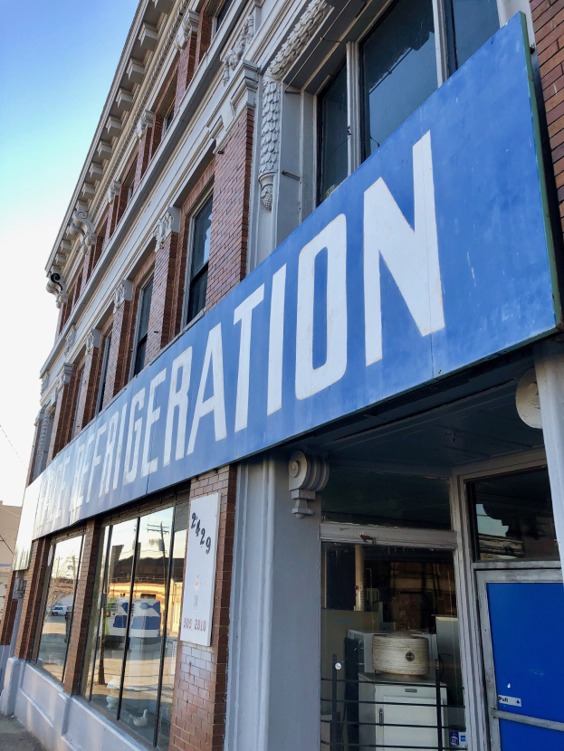 Außenansicht eines Kühlhauses mit Glaswänden, einer blauen Tafel und einem sichtbaren Kühlgerät im Hintergrund.