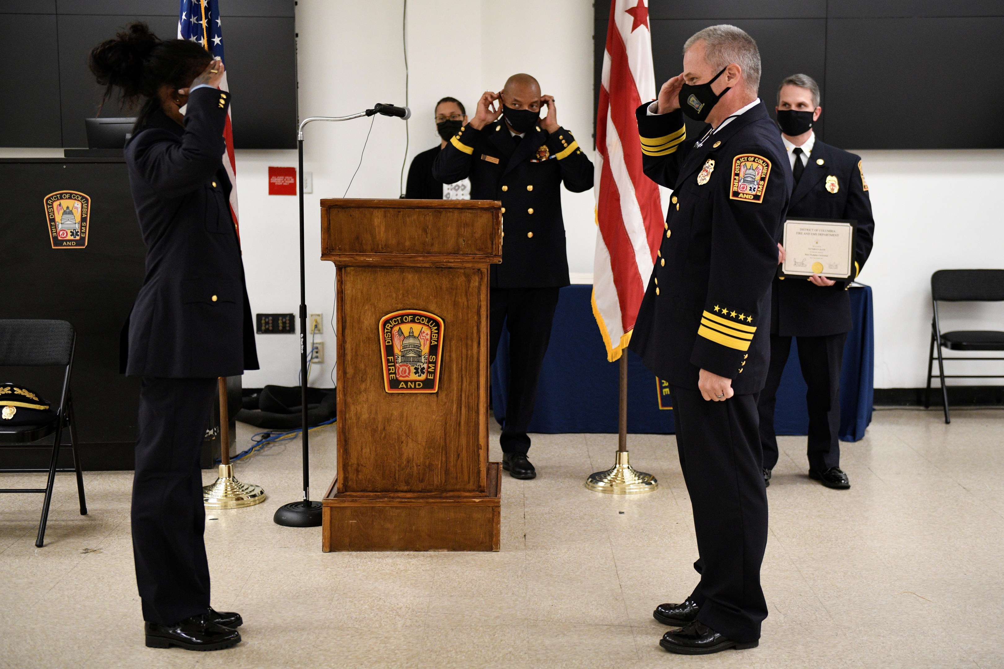 Gruppe uniformierter Personen in Masken vor einem Rednerpult bei einer Vereidigung, eine hält ein Zertifikat, mit Flaggen und Sitzplätzen im Hintergrund.