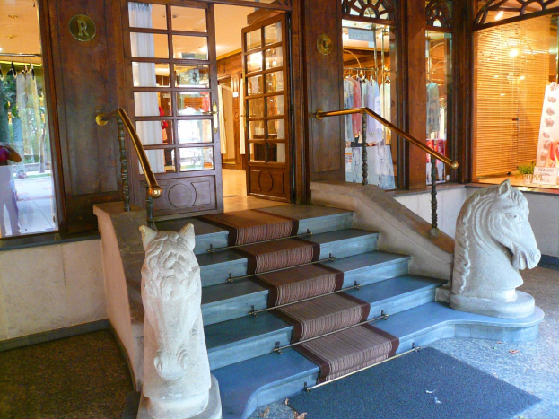 Eingang zu einem Geschäft mit zwei Pferdestatuen zu beiden Seiten der Treppe, Teppichboden, Glastüren, Schaufensterpuppen in Kleidern und dekorative Beleuchtung.