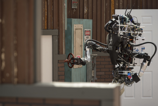 Ein humanoider Roboter mit ausgestreckten Armen und geneigtem Kopf steht vor einem Gebäude mit einer sichtbaren Tür und Wand dahinter.