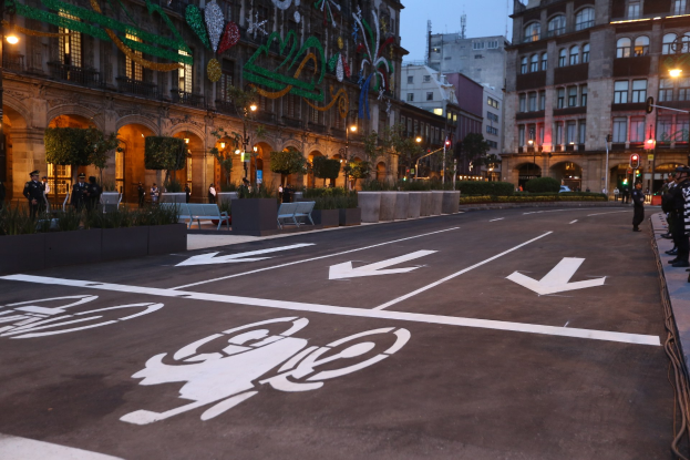 Stadtstraße mit einem ausgewiesenen Fahrradweg, umgeben von Gebäuden, Straßenmöblierung, Fußgängern auf dem Gehweg, Grünflächen und einem klaren blauen Himmel.