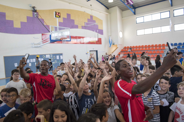 Kinder vor einem Basketballfeld mit Handys stehend, mit einer Anschlagtafel, Uhr, Torpfosten, Basketballnetz, Deckenleuchten, Stühlen und Fenstern im Hintergrund.