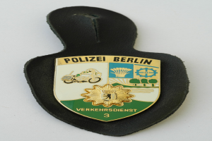 Ein schildförmiges Polizeisiegel mit "Polizei Berlin" in fetter schwarzer Schrift auf einem blauen Hintergrund mit einem weißen Stern und Rahmen, das auf einer Oberfläche ruht.