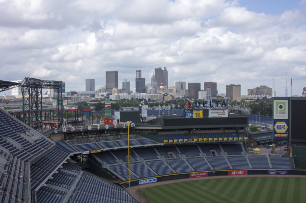 Ein Baseball-Stadion mit einer Stadtkulisse im Hintergrund, das leere Sitzplätze, Pfosten und Schilder zeigt, auf einem grasbewachsenen Feld mit fernen Bäumen und Gebäuden unter einem bewölkten Himmel.