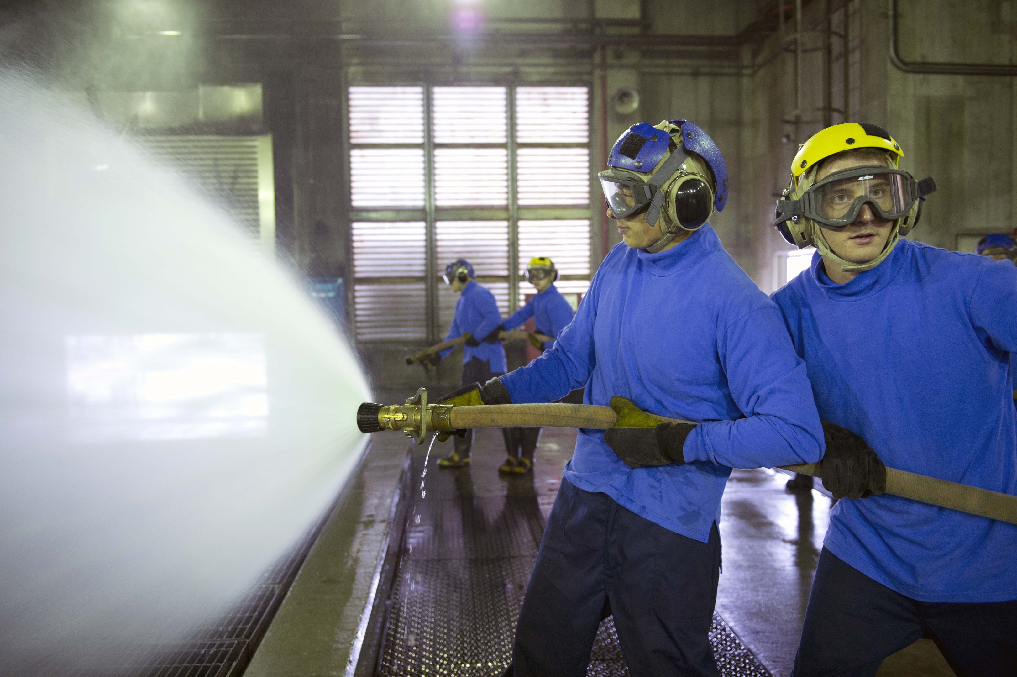Eine Gruppe von Männern in blauen Hemden und gelben Helmen arbeitet an Maschinen in einer Fabrik, wobei einer einen Schlauch hält und Wasser auf den Boden sprüht.