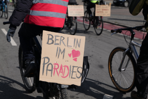 Eine Gruppe von Menschen, die auf Fahrrädern eine Straße entlangfahren, mit einem "Berlin I'm Paradies"-Schild im Vordergrund und einem Auto im Hintergrund, das Bild leicht unscharf.