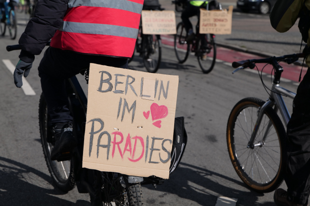 Eine Gruppe von Menschen, die auf Fahrrädern eine Straße entlangfahren, mit einem "Berlin I'm Paradies"-Schild im Vordergrund und einem Auto im Hintergrund, das Bild leicht unscharf.